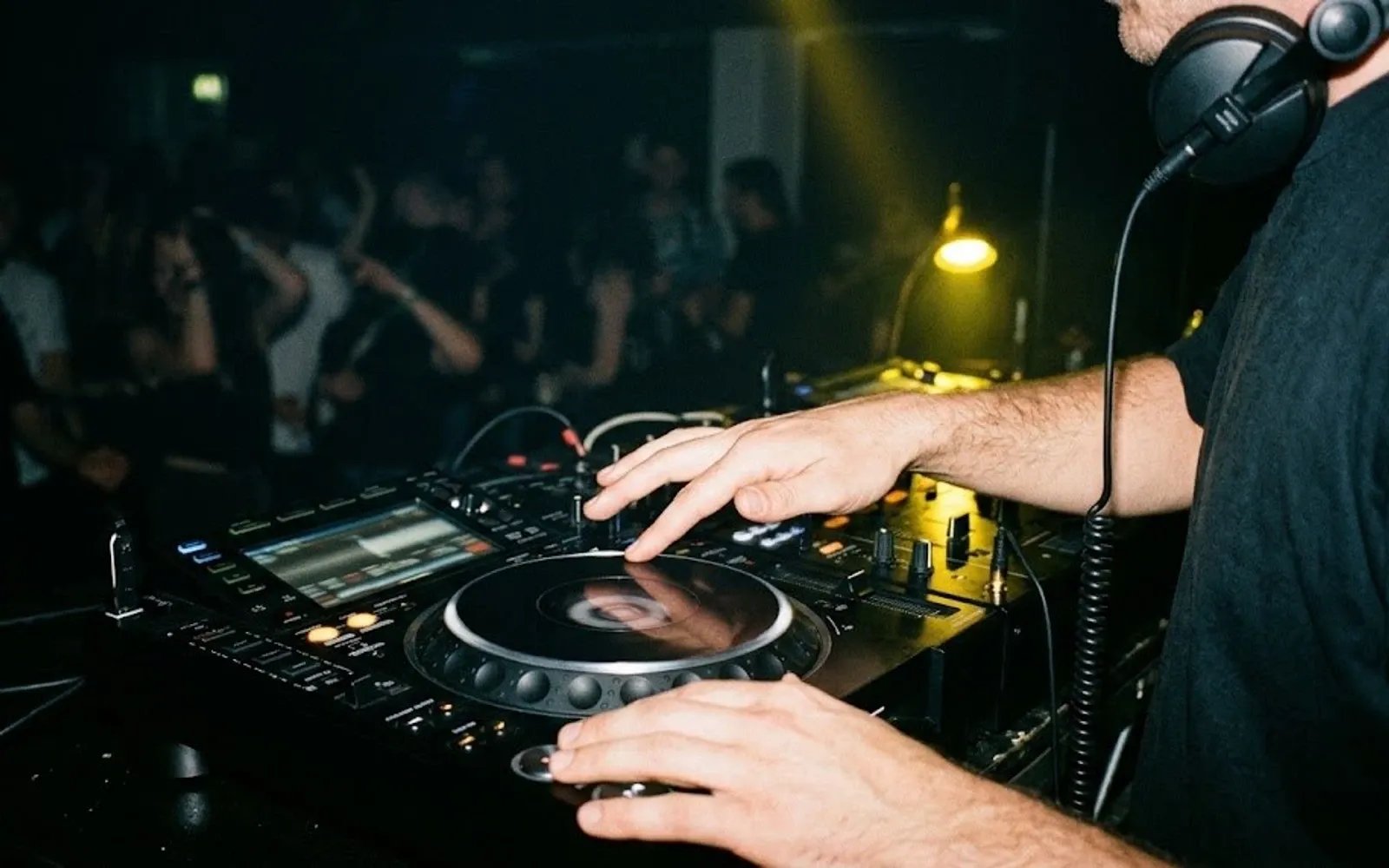 DJ ze słuchawkami koryguje tempo jogiem odtwarzacza, w tle tańczący tłum w żółtym świetle